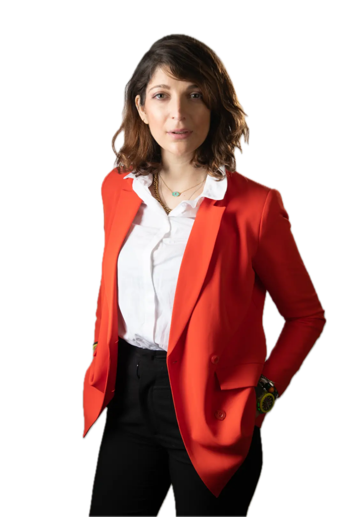Portrait professionnel d'une femme aux cheveux bruns ondulés, vêtue d'une chemise blanche et d'un blazer rouge vif, posant sur un fond uni beige clair.